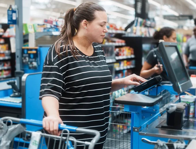 Woman in grocery store checkout line with one hand on grocery cart and other on electronic key pad next to conveyor belt