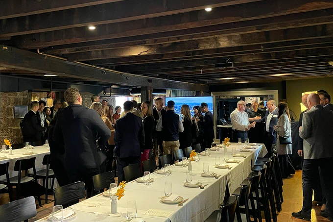 A group of people are gathered in a dimly lit, rustic restaurant with wooden beams and long tables set with white tablecloths and glasses. Yellow flowers are arranged as centrepieces. Some individuals are engaged in conversation near a window letting in daylight.