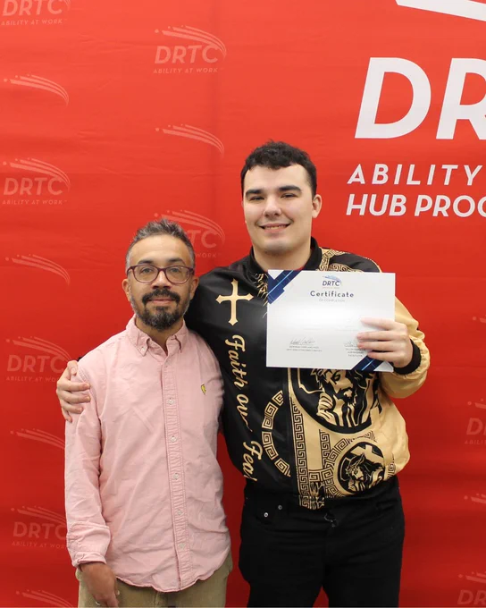 Two men stand in front of a red DRTC backdrop, one holding a certificate of achievement with a proud smile.