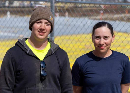 Two individuals stand side by side outdoors in front of a chain-link fence with a yellow field behind them on a sunny day.