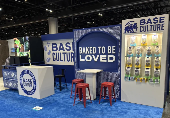 A visually appealing trade show booth for Base Culture with bright blue and red accents. It features product displays and the slogan 'Baked to be Loved.'