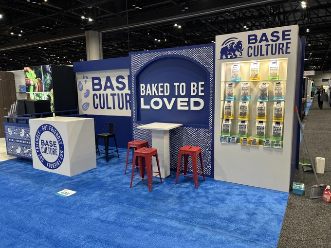 A trade show booth for Base Culture featuring product displays and branded signage. The setup includes a promotional message 'Baked to be Loved' emphasizing their bakery products.