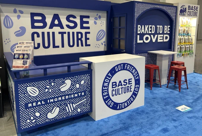 A bright, engaging trade show booth for Base Culture featuring bold blue signage. The booth highlights gut-friendly products and real ingredients, with stools for visitors.