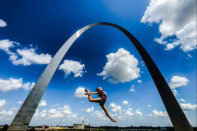 A woman leaps and stretches her foot behind her head under the Gateway Arch.