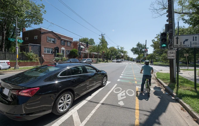 Photo of a man on a bicycle stopped in the bike lane next to a black car on the road.