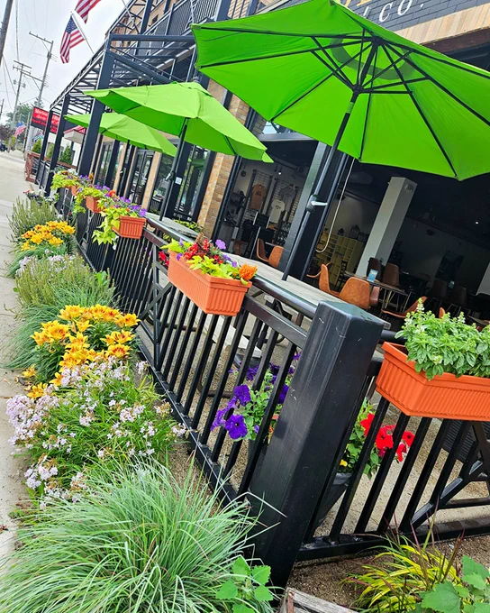 A sidewalk view of an outdoor patio area with green umbrellas and orange planters attached to a black fence. The planters are filled with vibrant flowers in shades of yellow, orange, and purple. An American flag is visible in the background.