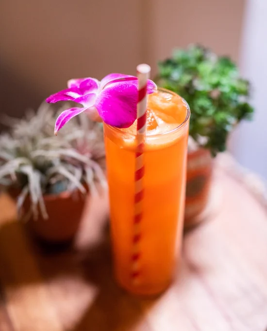 A tall glass of orange juice adorned with a purple orchid and a red-and-white striped straw. The drink is placed on a wooden surface with two potted plants in the background.