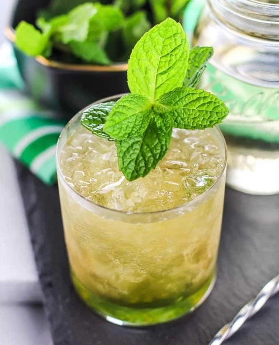 A glass of mint julep is on display with crushed ice, garnished with fresh mint leaves. A jar is visible in the background, and a bowl of mint is partially shown.
