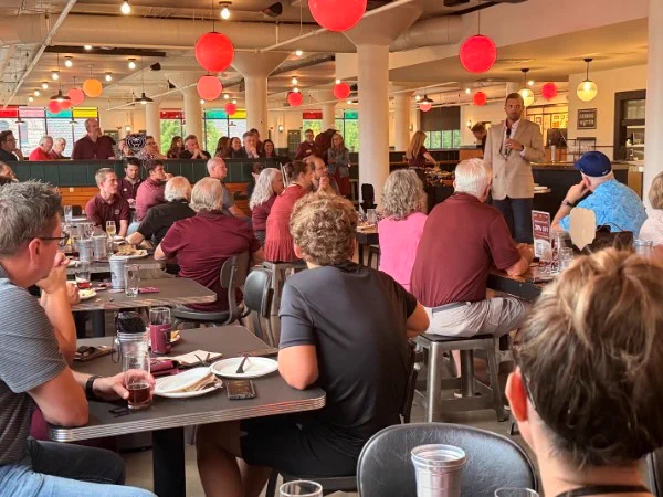 A group of people seated in a restaurant with red and orange lanterns overhead, listening to a speaker standing and holding a microphone. The room is lively and filled with attentive attendees.