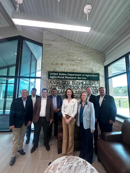 A group of people is standing in an indoor space near a sign that reads "United States Department of Agriculture Agricultural Research Service, Knipling-Bushland U.S. Livestock Insects Research Laboratory." They are dressed in business attire, with large windows in the background.