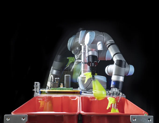 Motion-blurred robot moves a green spray bottle between red containers.