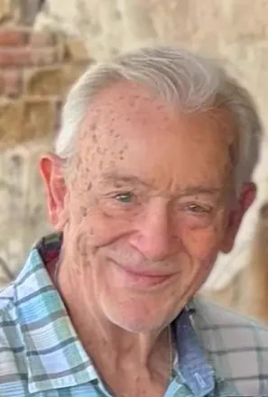 Elderly man with white hair wearing a blue and green plaid shirt standing in front of a rustic brick wall.