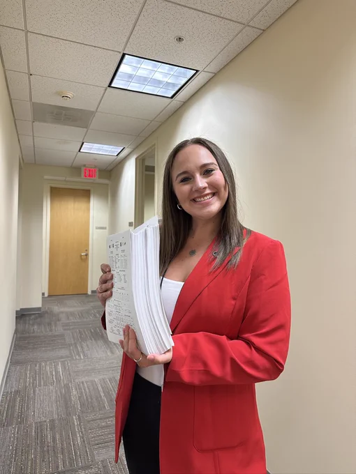Natalie Gore wearing a red blazer and holding a large stack of papers in a brightly lit office hallway.