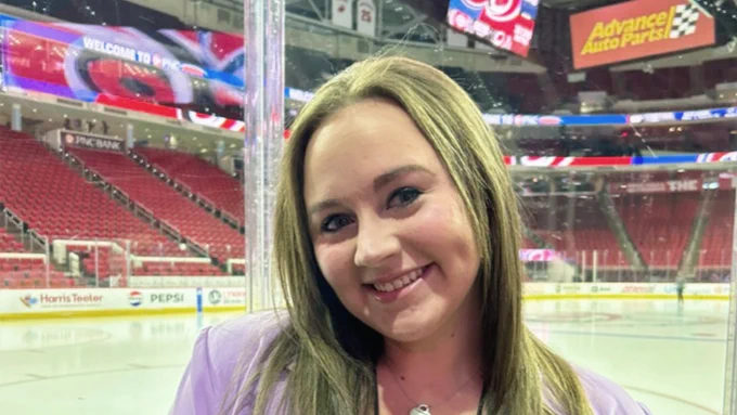 Woman in a lavender blazer standing inside an empty ice hockey arena near the glass barrier.
