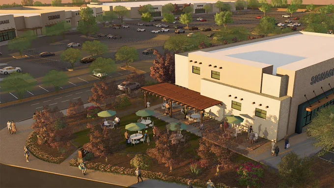This image shows a render of a shopping area with multiple buildings. The scene includes parking lots with cars and people walking. In the foreground, there's an outdoor seating area with tables, chairs, and umbrellas surrounded by landscaping.