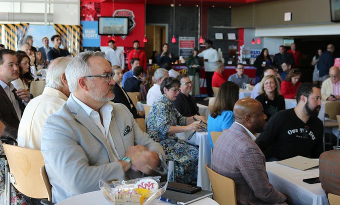 A group of people seated at tables are attentively listening at an indoor event. Some individuals are holding drinks or taking notes. The atmosphere appears engaged and focused.