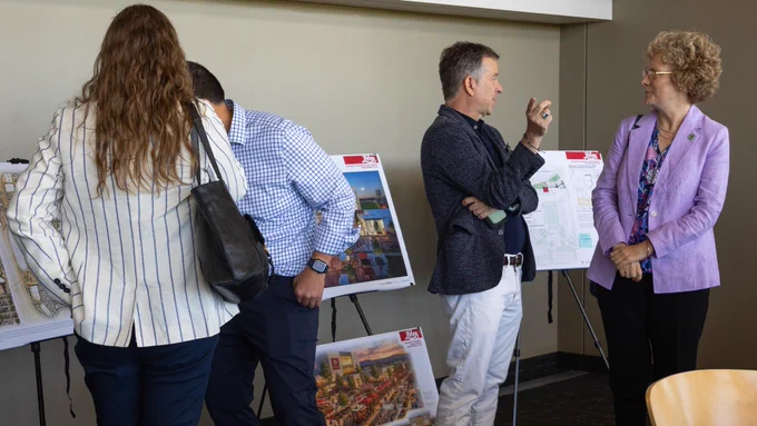 A group of people is gathered around display boards on easels, each showing urban planning images and maps. Two individuals in the foreground are engaged in conversation, while others examine the displays.