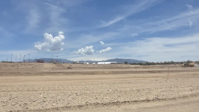 A vast, dry construction site under a blue sky with scattered clouds. Mountains are visible in the distance. There is sparse vegetation around the site, and a few construction vehicles can be seen on the right.