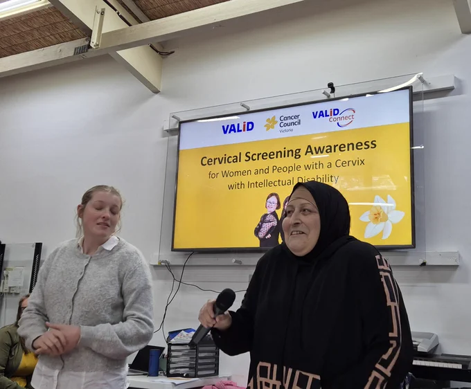 Two individuals present in front of a screen displaying a cervical screening awareness message for women and people with a cervix with intellectual disability.