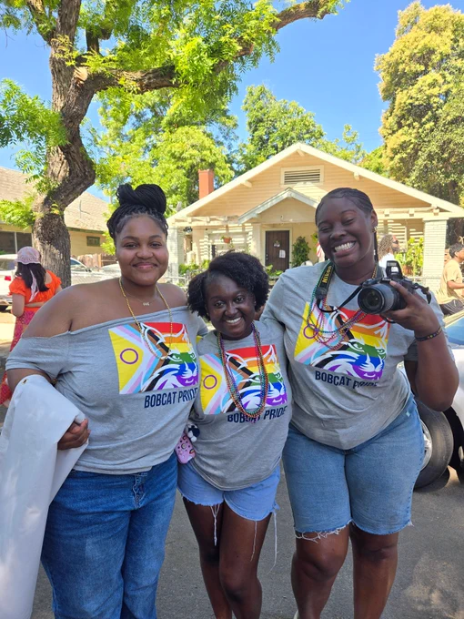 Three friends wearing matching gray Bobcat Pride t-shirts stand together outdoors as the prepare to walk in Fresno's Pride parade.