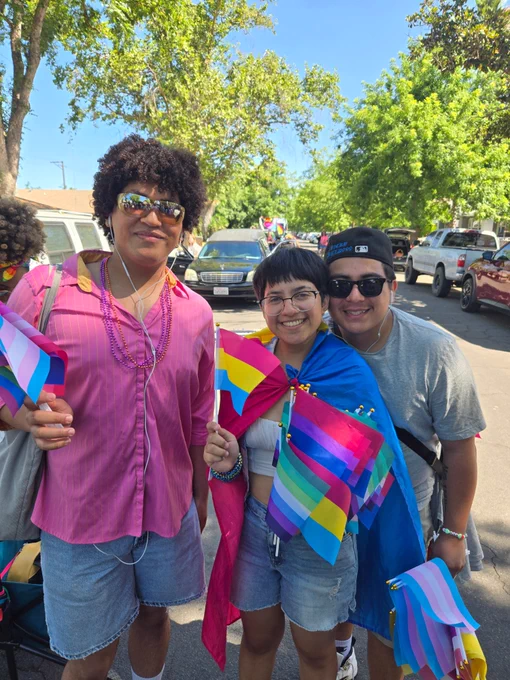 Three students stand outdoors holding and wearing colorful pride flags as they prepare to walk in Fresno's Pride parade.