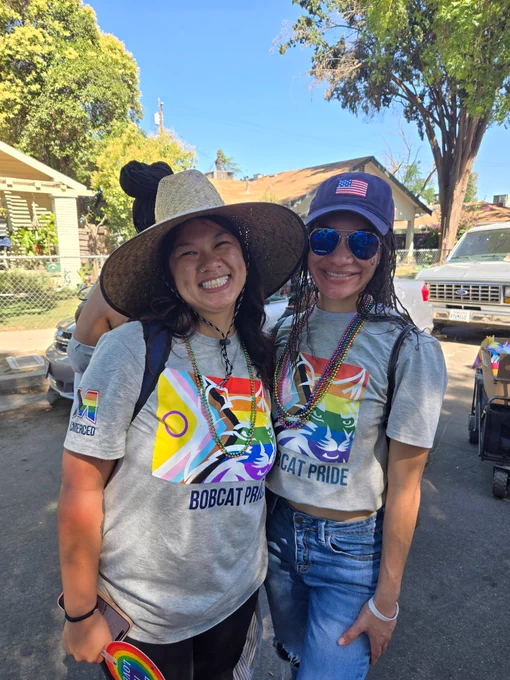 Two students wearing gray Bobcat Pride t-shirts with rainbow designs and colorful bead necklaces standing outdoors on a sunny day.