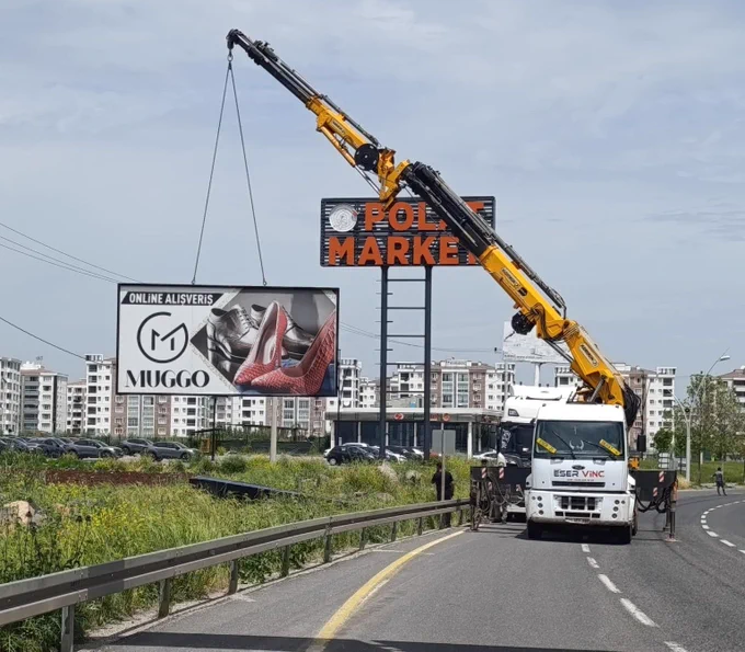 Fotoğrafta geniş bir bulvar kenarında gerçekleştirilen reklam panosu kaldırma çalışması görülüyor. Yol üzerine konuşlandırılmış büyük bir vinç, metal halatlar yardımıyla büyük boyutlu bir reklam panosunu yerinden söküyor. Panoda ayakkabı reklamı yer alırken, pano havada asılı halde taşınmaya hazırlanıyor.

Vinç aracının yanında çalışma yürüten görevliler bulunuyor. Arka planda alışveriş merkezi ve çok katlı konut blokları görülürken, yol kenarında bariyerler ve yeşil alanlar yer alıyor. Gökyüzü bulutlu bir görünüme sahip ve çalışma açık alanda gerçekleştiriliyor.

Fotoğraf, kent estetiğini ve trafik güvenliğini olumsuz etkilediği değerlendirilen reklam yapılarının kaldırılmasına yönelik belediye çalışmasını belgelemektedir. Sahne, mevzuata aykırı reklam panolarının vinç desteğiyle sökülerek kaldırıldığı bir uygulamayı yansıtmaktadır.