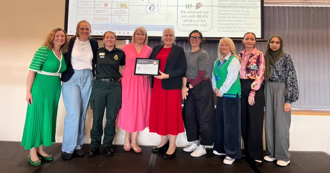 A group of nine people standing together on a stage, smiling. One person in the centre is holding a certificate. They are in front of a screen displaying text and graphics related to a maternity tool achievement.