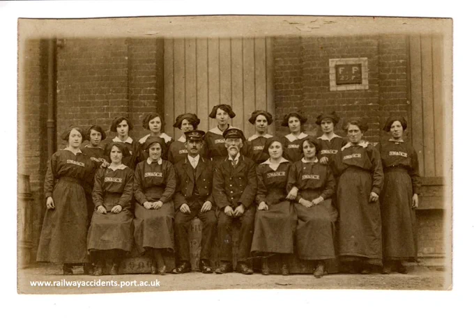 Photographic postcard, showing a group of 15 railwaywomen arranged around 2 railwaymen in the centre. In the front row two women sit either side of the men; behind them 11 women stand. All are in uniform; the women are carriage cleaners, and have 'SW & SCR' stitched in. Several of the women smile; at least 2 have what appear to be National Union of Railwaymen lapel badges; most are young, several are somewhat older. They are arranged in front of a brick-built  goods shed, with two sliding wooden doors closed.