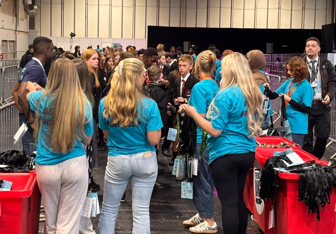 A group of people stand in a large indoor venue. Several individuals are wearing blue shirts and distributing lanyards from red bins. A crowd gathers in front of them, waiting in line. The space is busy and bustling, with various attendees holding event passes.