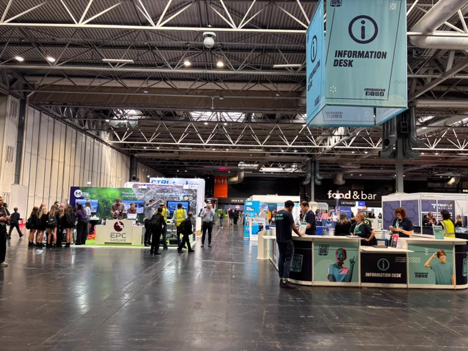 The image shows a bustling exhibition hall with various booths and information desks. People are gathered around booths, engaging and walking through the space. Overhead, there's a large sign for an information desk.