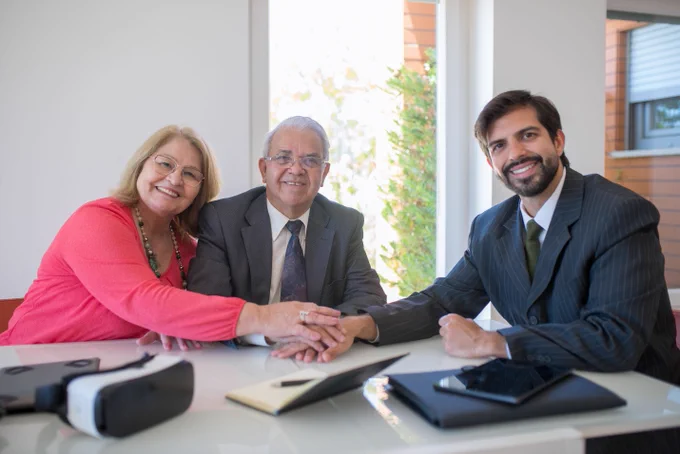 Tres personas, una mujer y dos hombres sentados en una mesa vestidos de traje dándose la mano en señal de acuerdo.