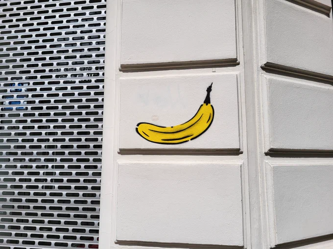 Zu sehen ist eine Nahaufnahme einer Häuserecke mit großen, hellen Steinen. Auf einen davon ist eine Banane gemalt. 
Links daneben ist ein heruntergelassener Rollladen zu sehen, wohl vor einem Ladengeschäft.