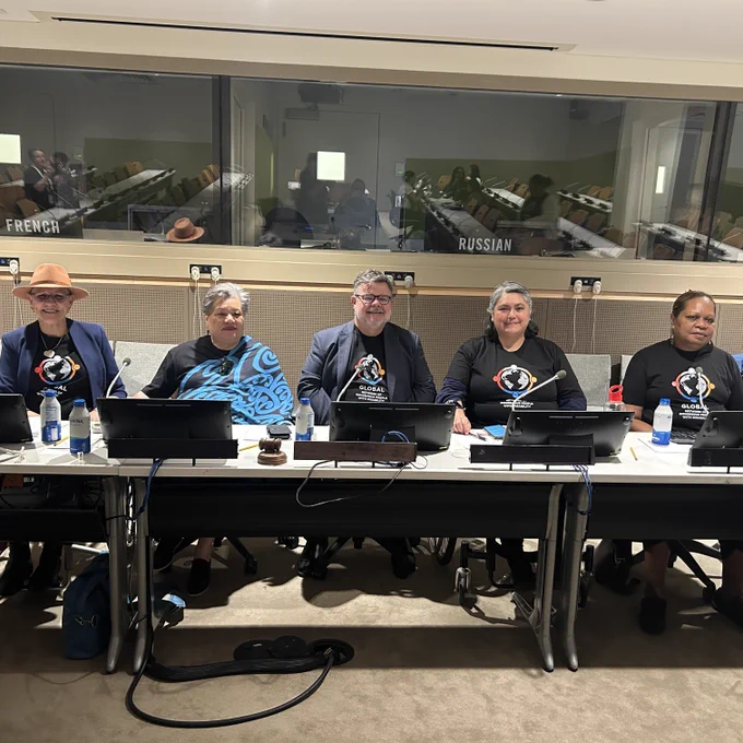 A photograph of five people seated panel-style behind a long white desk inside an international conference room or United Nations chamber. Most panel members are wearing matching black t-shirts featuring the logo for the Global Network of Indigenous People with Disability. The desk in front of them is equipped with microphones, laptops, water bottles, and a wooden gavel.