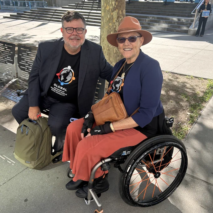 A sunny outdoor photograph of FPDN CEO Damian Griffis and Chairperson Aunty Kay Sadler smiling together on a paved walkway. Damian is seated on a bench on the left, wearing a black t-shirt with the Global Network of Indigenous People with Disability logo. Aunty Kay Sadler is positioned on the right, using a manual wheelchair. She wears a matching network t-shirt.