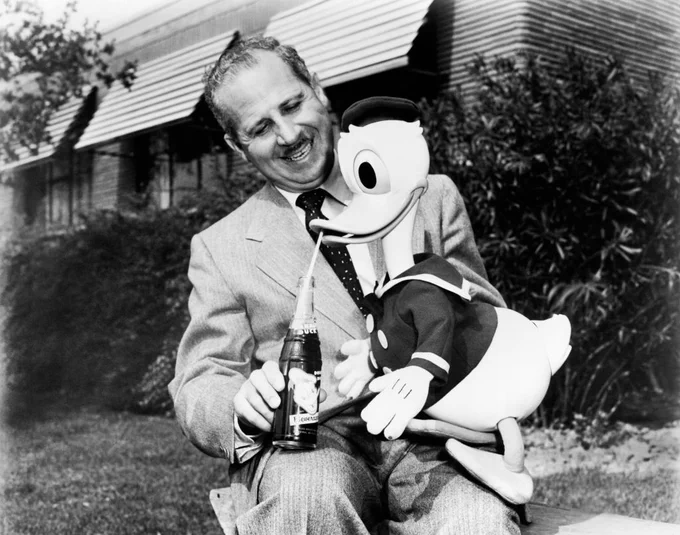 A person smiling while sitting on a bench outdoors, holding a bottle of Coca-Cola. Next to them is a large Donald Duck figure also holding a Coke bottle.