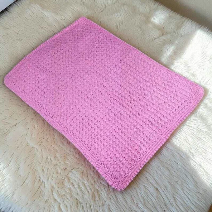 A rectangular bright pink crocheted blanket is laid flat, showcasing a dense, textured pattern across its surface. It rests on a soft, cream-colored shaggy rug. Sunlight streams across, casting gentle shadows and highlighting the texture, creating a warm and cosy atmosphere.