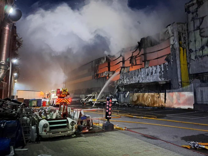 Photo at night showing a hose being used to extinguish a fire at a warehouse unit. A plume of smoke in the background.