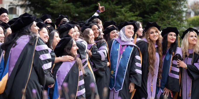Group of graduates in black and purple academic robes posing together outdoors for a celebratory photo.