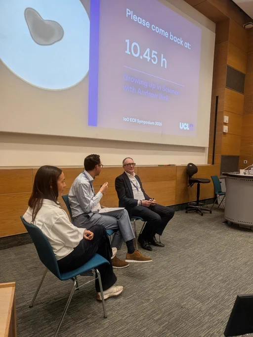 Three seated speakers take part in a discussion panel in a lecture theatre, with a projected slide behind them reading “Please come back at 10.45 h – Growing Up in Science with Andrew Dick, IoO ECR Symposium 2026, UCL.”
