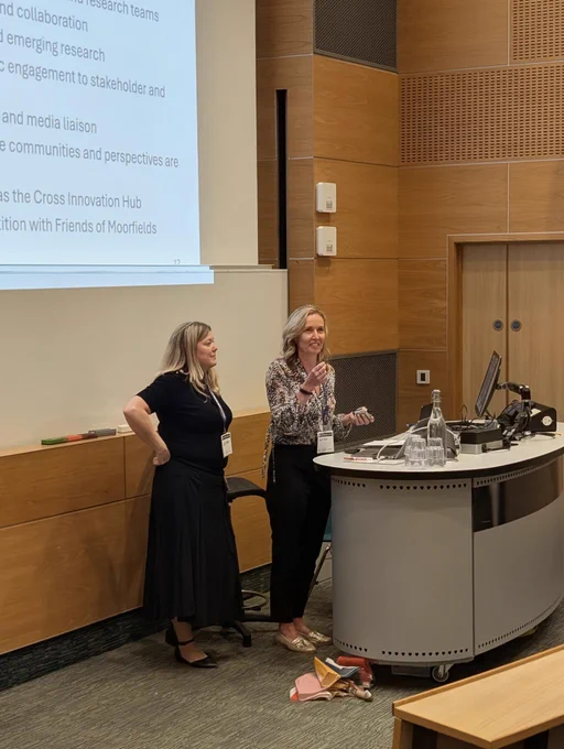 Two speakers stand at the front of a lecture theatre beside a podium and computer, presenting in front of a projected slide with bullet points; glasses and a water bottle sit on the desk.