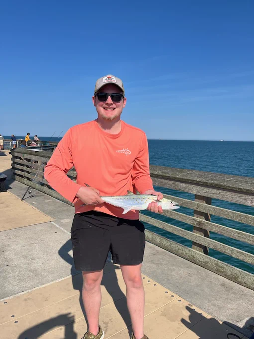 Jayden stands on Jennette's Pier holding a Spanish mackerel while wearing a bright orange shirt, shorts, and a cap. The ocean and blue sky are in the background, with fishing rods visible along the railing.