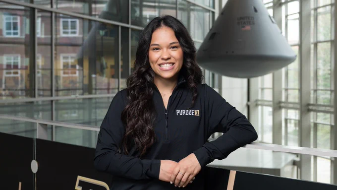 Abigail Mizzi smiles in Neil Armstrong Hall of Engineering wearing a Purdue pullover, with a space capsule visible in the background. 