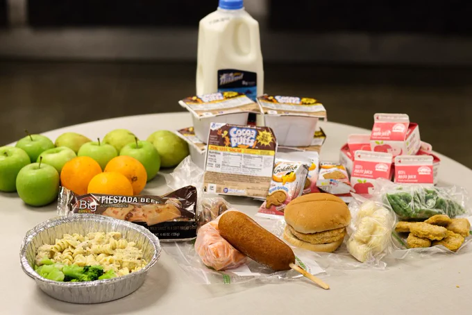 A table displays the full contents of a weekly meal pack, including fresh apples and oranges, a half‑gallon of milk, multiple cartons of apple juice, single‑serve Cocoa Puffs cereal cups, packets of Giant Goldfish cinnamon graham crackers, a foil tray of chicken alfredo with broccoli, a packaged turkey corn dog with carrots, chicken nuggets with green beans, a chicken sandwich with tater tots, and a Fiestada beef stuffed sandwich with refried beans. The items are arranged neatly to show the variety included in the free breakfast and lunch packs for children.