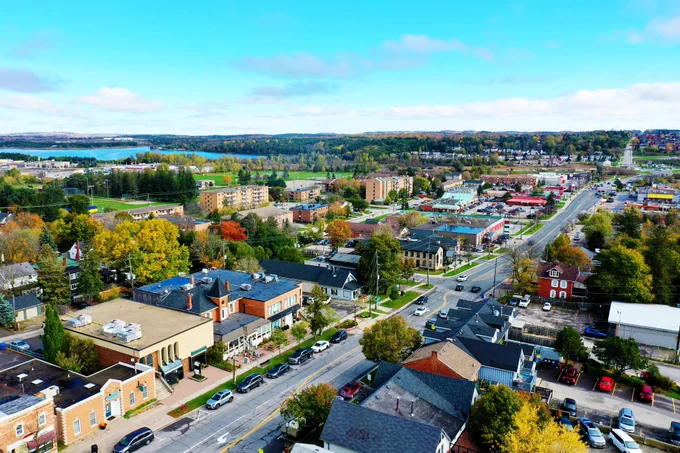 Aerial view of Orangeville, showing a main road lined with buildings, homes, trees, and parked cars, with a lake and surrounding greenery visible in the distance under a bright blue sky.