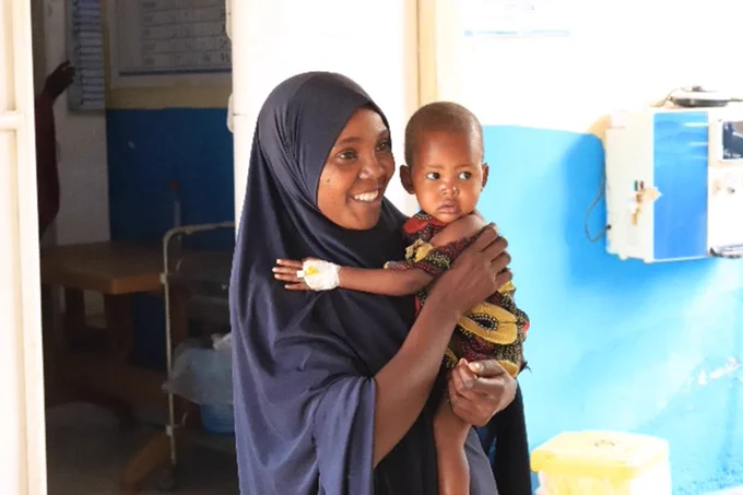 Woman in a dark hijab holding a young child with a medical bandage on their hand inside a healthcare facility in Somalia.