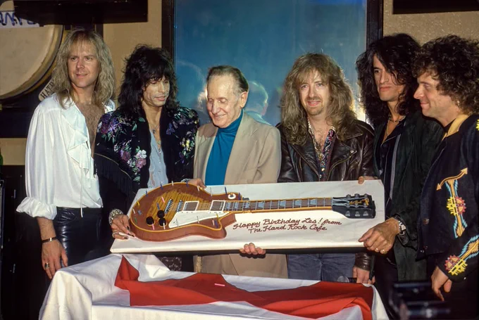Aerosmith and Les Paul stand together, holding a large decorated Gibson Les Paul guitar-shaped cake in a room. The cake has a message wishing a happy birthday. There is some text on the cake, indicating it's from the Hard Rock Cafe. The setting suggests a celebration or event.