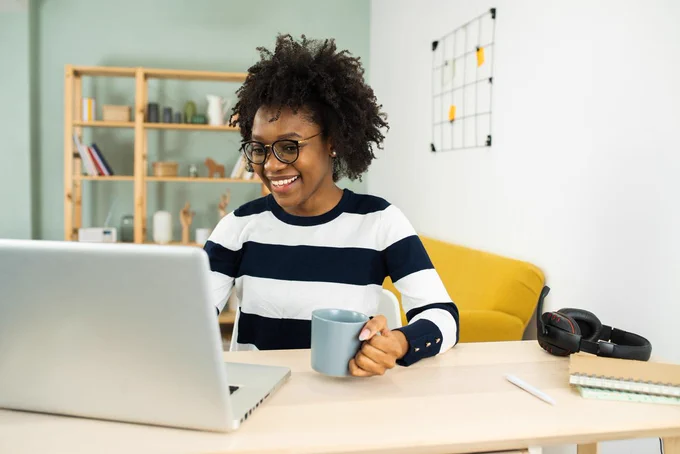 A Black woman smiles while looking at a laptop screen, holding a mug, seated at a desk with headphones and a notebook nearby in a bright home workspace.