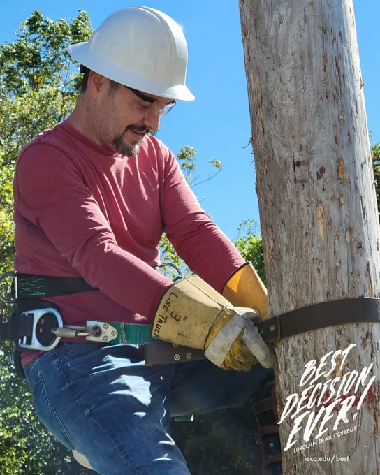 Utility worker wearing a white hard hat and gloves securing a strap around a wooden utility pole on a clear day.
