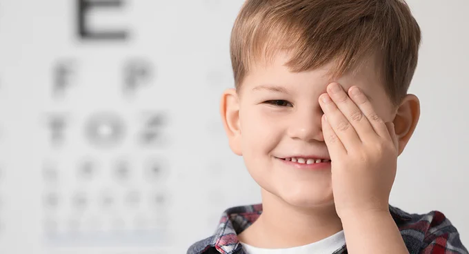 Child covering one eye during an eye exam with a blurred face and a Snellen chart in the background.